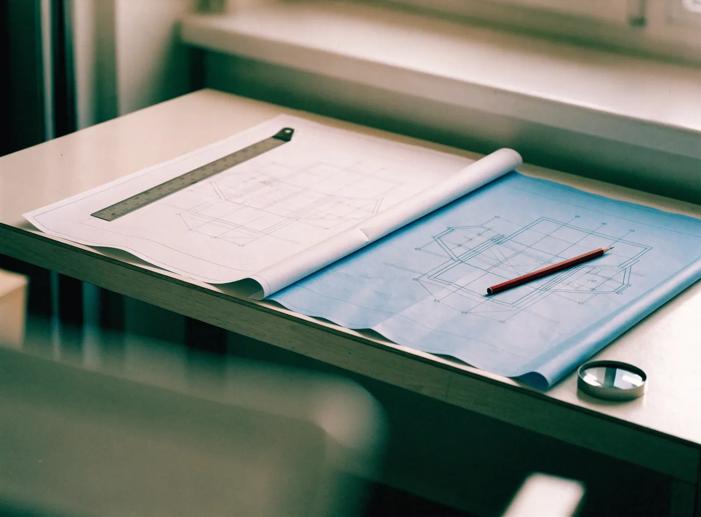 Two construction drawing sheets laid side by side on a desk for comparison, a metal scale ruler across one and a red pencil on the other